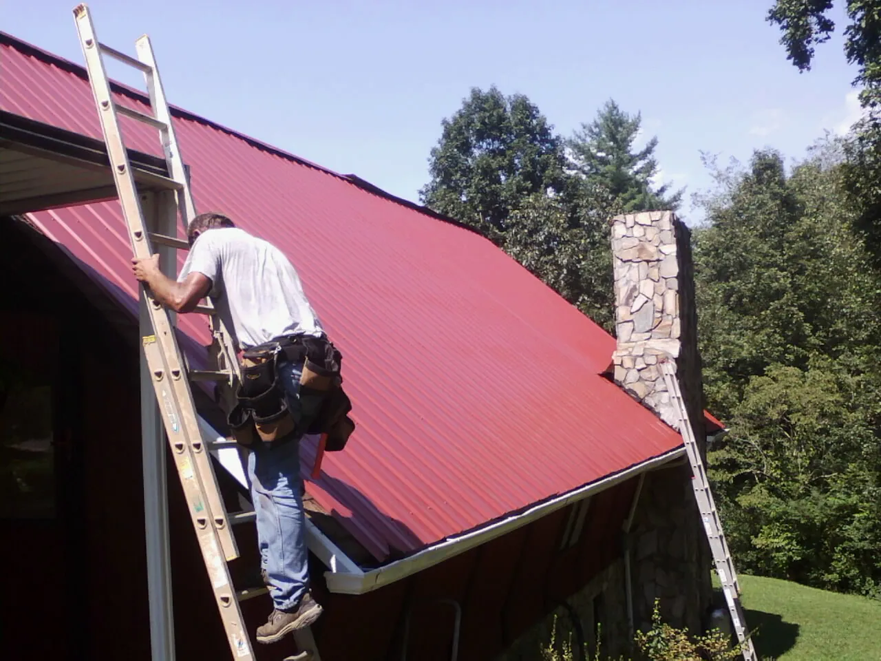 Roofer on a ladder beside a red metal roof and stone chimney — Soffit & Fascia Repair by Roofing Brothers in Waukegan